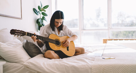 Brunette female learning how to play guitar while watching online video course on laptop computer in cozy home interior. Young woman with netbook enjoying hobby with musical instrument relaxing on bed - obrazy, fototapety, plakaty