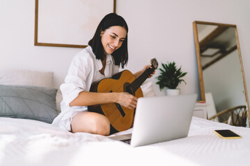 Smiling female learning how to play guitar while watching online video course on laptop computer in cozy home interior. Young woman with netbook enjoying hobby with musical instrument relaxing on bed