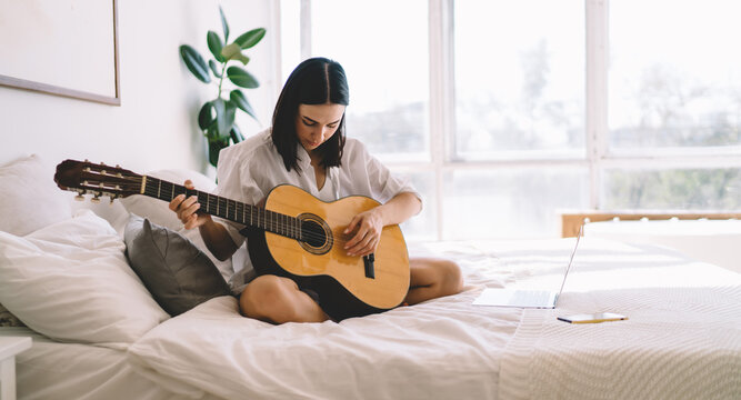Brunette female learning how to play guitar while watching online video course on laptop computer in cozy home interior. Young woman with netbook enjoying hobby with musical instrument relaxing on bed