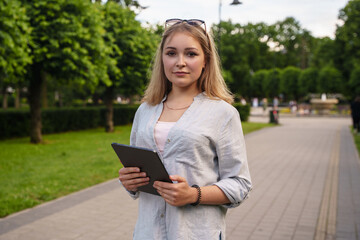 Portrait of young female student holding tablet in summer public park in sunny day.
