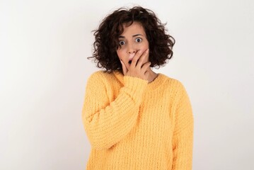 young beautiful woman with curly short hair wearing yellow sweater over white background covers mouth and looks with wonder at camera, cannot believe unexpected rumors.