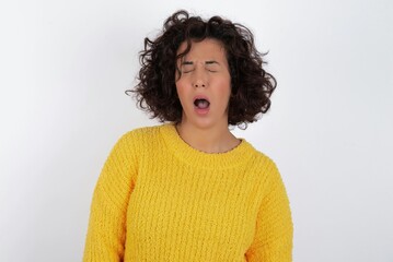 young beautiful woman with curly short hair wearing yellow sweater over white background yawns with opened mouth stands. Daily morning routine