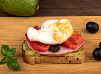 Sandwich with avocado, slices of ham and tomato with poached egg on a cutting board.