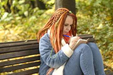 Young woman student with long hair in casual clothes sitting on wooden bench and writing notes in notebook