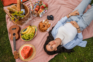 Shot of cheerful young woman at picnic lying on blanket around food and drink in park.