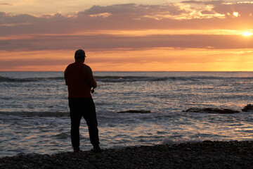 A lonely figure of a fisherman with a fishing rod in his hand on the seashore at sunset. Contour...