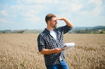 Happy farmer proudly standing in a field. Combine harvester driver going to crop rich wheat harvest. Agronomist wearing flannel shirt, looking at camera on a farmland