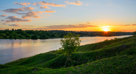 Summer sunny landscape with sun rising over the blue calm river and green hills.