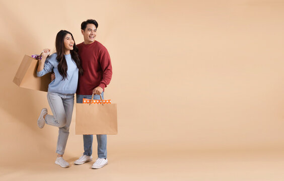 Happy smiling Asian couple with shopping bags in sweater enjoying their winter discount sale isolated on beige color background. Shopaholic fashionable 20s man and woman looking away with copy space.