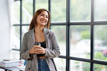 Beautiful Asian businesswoman standing with coffee to at work and think of new projects in the office.