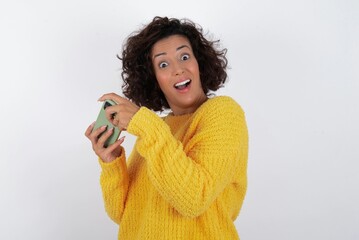 Nice addicted cheerful young beautiful woman with curly short hair wearing yellow sweater over white background using gadget playing network game