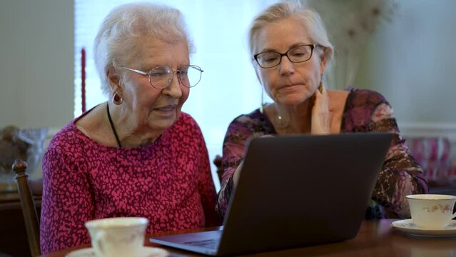Happy elderly middle aged woman sitting at table with grown up daughter, watching family or journey photos on computer. Smiling blonde lady listening to older, using laptop.