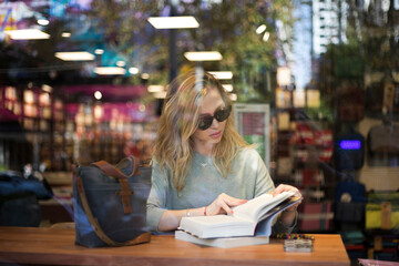 View through the book shop window of young blonde woman reading book, wearing sunglasses. 