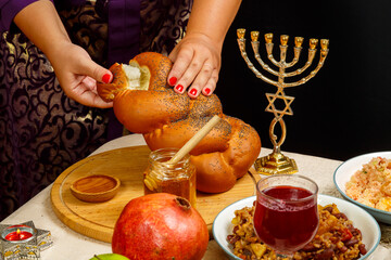 A woman's hand breaks challah on a wooden board on the holiday of Rosh Hashanah near honey, apples and menorah.