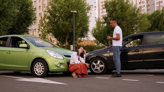 A man shouts at an upset woman near two wrecked cars after an accident on the road. A guilty woman is crying sitting on the ground near a broken car. Car accident.