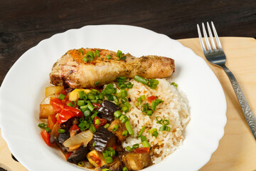 Rice with vegetables in sauce and chicken leg in a white plate on a handmade wooden board next to a fork on a wooden table.
