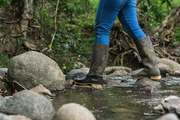 migrant woman crossing a stream wearing blue jeans and black marsh boots. hiking through the dense...