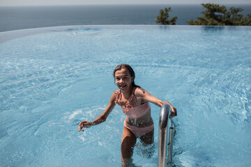 A young teenager girl in a swimsuit, swimming in a blue pool against the background of the sea and sky. The female is out of the pool