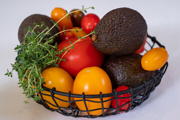 black metal basket with colorful vegetables, red and yellow tomatoes, avocado and green thyme on white kitchen table