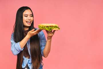 Young beautiful asian japanese chinese woman eating sandwich or big burger with satisfaction. Girl enjoys tasty hamburger takeaway, diet concept, standing isolated over pink background.