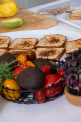 black metal basket with colorful vegetables, red and yellow tomatoes, avocado and green thyme and appetizing toasts on white kitchen table