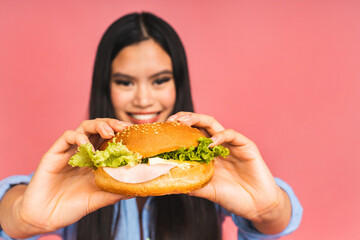 Young beautiful asian japanese chinese woman eating sandwich or big burger with satisfaction. Girl enjoys tasty hamburger takeaway, diet concept, standing isolated over pink background.