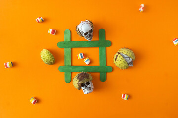 A tic tac toe game made with green sticks, candies and sculls in chestnut shells on orange background. Surreal concept for Haloween or Autumn banner or advertisement. Flat lay - obrazy, fototapety, plakaty
