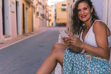 A brunette woman sitting smiling at the camera while holding her mobile phone with both hands in the colorful streets of Villajoyosa, Alicante, Spain.