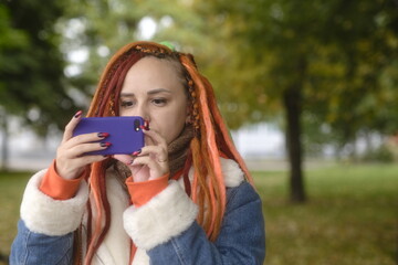 Young pretty woman with dreadlocks photographing on mobile phone, standing in parkland. Female tourist with colourful multicoloured hairstyle taking pictures on gadget.