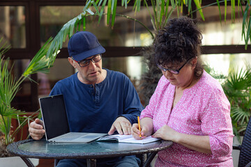 Mature man and woman in glasses talking to each other, sitting at table, write in notepad near...