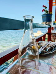 an empty bottle of wine is on the table, next to an iron plate containing a used fork and knife, as well as a slice of lemon and a used napkin. outside, the sea splashes nearby