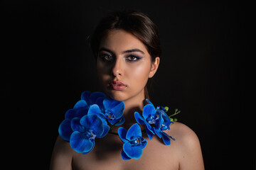 Gorgeous, romantic serious brunette woman with blue make up on perfect skin and blue flowers on neck on black background