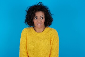 young beautiful woman with curly short hair wearing yellow sweater over blue wall with thoughtful...
