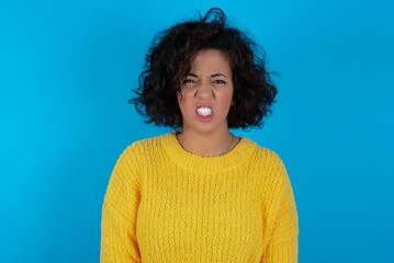 Mad crazy young beautiful woman with curly short hair wearing yellow sweater over blue wall clenches teeth angrily, being annoyed with coming noise. Negative feeling concept.