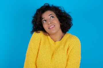 Portrait of mysterious young beautiful woman with curly short hair wearing yellow sweater over blue wall looking up with enigmatic smile. Advertisement concept.