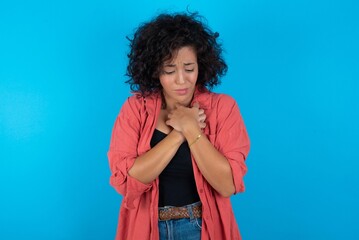 Fototapeta na wymiar Sad young beautiful woman with curly short hair wearing red overshirt over blue wall feeling upset while spending time at home alone staring at camera with unhappy or regretful look.