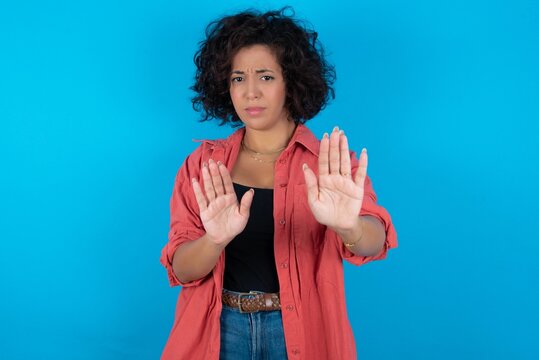 Afraid young beautiful woman with curly short hair wearing red overshirt over blue wall, makes terrified expression and stop gesture with both hands saying: Stay there. Panic concept.