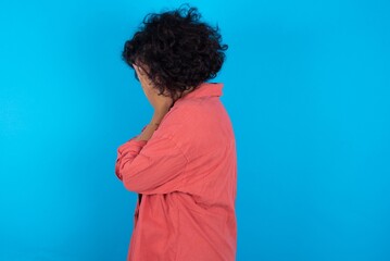 Sad young beautiful woman with curly short hair wearing red overshirt covering face with hands and...