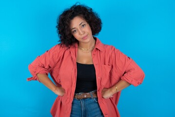 Funny frustrated young beautiful woman with curly short hair wearing red overshirt holding hands on waist and silly looking at awkward situation.