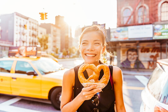New York people lifestyle photo. Woman eating pretzel in Manhattan, a classic New York City snack. Multiracial asian young professional portrait smiling at camera