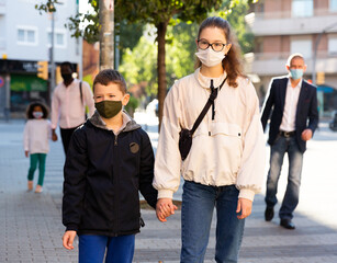 Teen girl wearing protective mask holding hand of her preteen brother walking along city street on warm autumn day. Concept of necessary precautions in COVID pandemic