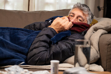 Homme malade allongé sur un canapé avec une écharpe et une couverture. Devant lui sur la table il y a un thermomètre et des médicaments