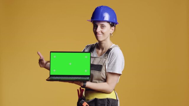 Female construction worker holding laptop with greenscreen in studio, pointing at chroma key display with blank copyspace. Using isolated mockup template on computer, repair work.