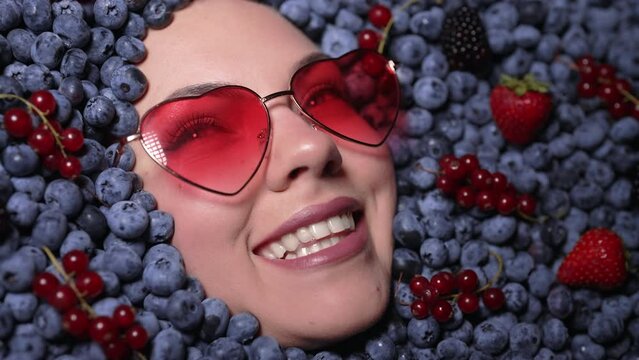 Happy woman face in eyewear fresh ripe berries - bilberries, strawberries, currant. Young girl covered with blueberries. Lady enjoying organic plant. Diet, antioxidant, healthy vegan food.
