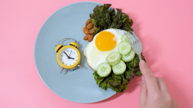 Hand puts an alarm clock on plate of food on pink background, concept of intermittent fasting.