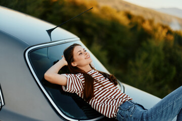 The woman driver stopped on the road and lay down on the car to rest and look at the beautiful landscape in a striped T-shirt and jeans. Complicated journey to nature