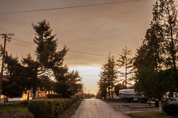 Fototapeta na wymiar Typical countryside road in the village of Banatsko novo selo, a serbian village of the Banat region of Vojvodina, Serbia, at dusk with an old rusted trucks parked, waiting to plough fields.....
