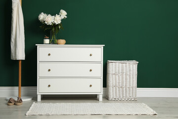 Modern white chest of drawers, coat stand and wicker basket near green wall indoors