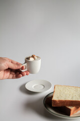 Hand holding cup of coffee with marshmallow and slice bread on white background. Morning breakfast, coffee break. Tasty beverage and sweet bakery. Winter and Autumn  concept