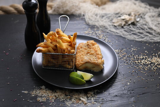 Fried breaded fish fillet served with potato fries in a metal serving basket, on black plate, selective focus.